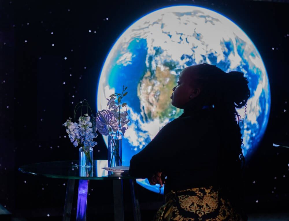 Naom Sang wearing a black, long sleeved top and yellow and black  pants, leans on a glass table with floral arrangements, thoughtfully facing a large digital display of Earth